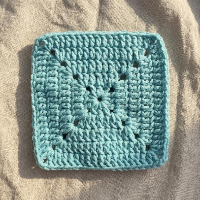 Peut inclure: Un carr&eacute; granny crochet bleu clair. Le carr&eacute; fait main pr&eacute;sente un motif de point textur&eacute; avec des coins ajour&eacute;s. Le carr&eacute; est pr&eacute;sent&eacute; sur un fond de tissu de couleur neutre, mettant en valeur la texture et la couleur du fil.