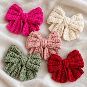 May include: Five handmade crochet bows in vibrant colors including hot pink, cream, dusty rose, sage green, and deep red. Each bow features a ribbed texture and a central wrapped knot detail, arranged in a cluster on a textured white fabric surface.