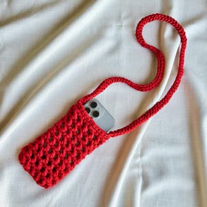Op de afbeelding: Een felrode gehaakte crossbody telefoontas met een open breistructuur en een lange, bijpassende rode riem. Een zilveren smartphone met een camera met drie lenzen is gedeeltelijk in de opening van het rechthoekige tasje gestoken.