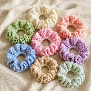 May include: A collection of handmade crochet scrunchies in various pastel colors, including light blue, pink, peach, lavender, green, and beige. The scrunchies are arranged on a neutral-colored fabric, showcasing their textured, ruffled design.