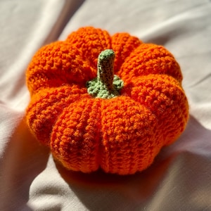 May include: A round, crocheted pumpkin crafted from bright orange yarn with a textured, ribbed surface. The top of the pumpkin features a small, light green crocheted stem and a ruffled base that connects the stem to the main orange body.