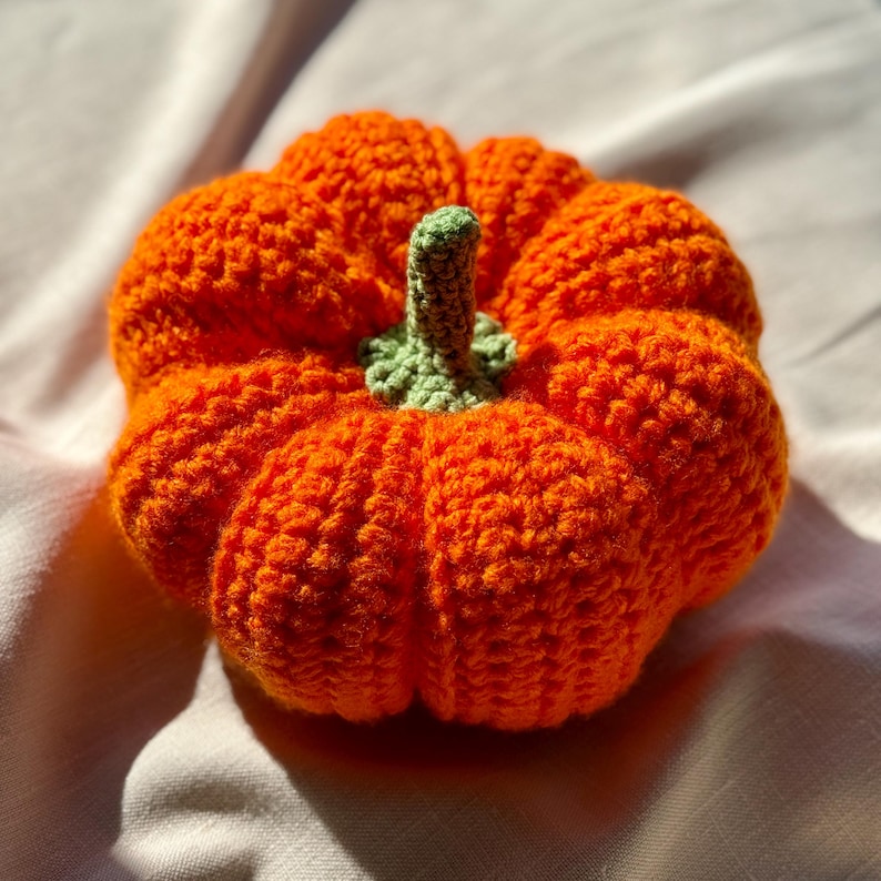 May include: A round, crocheted pumpkin crafted from bright orange yarn with a textured, ribbed surface. The top of the pumpkin features a small, light green crocheted stem and a ruffled base that connects the stem to the main orange body.