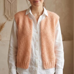 May include: A peach-colored, open-front knitted vest. The vest is made of a soft, textured yarn and has a simple, classic design. The vest is worn over a white button-down shirt.