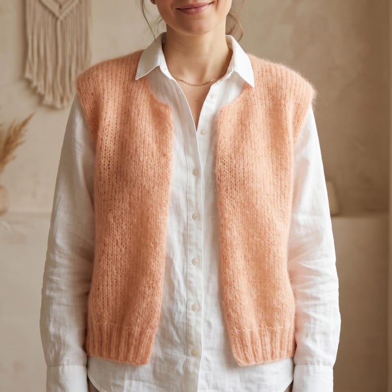 May include: A peach-colored, open-front knitted vest. The vest is made of a soft, textured yarn and has a simple, classic design. The vest is worn over a white button-down shirt.
