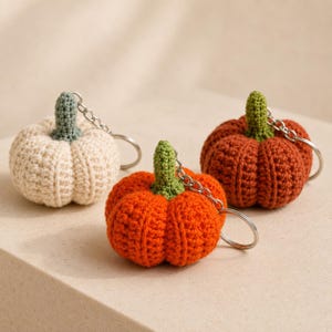 May include: Three crocheted pumpkin keychains featuring textured yarn in cream, bright orange, and burnt orange. Each pumpkin has a small green crocheted stem and is attached to a silver-toned metal split ring for keys or bag attachment.