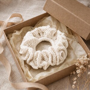 May include: A cream-colored, crocheted scrunchie sits inside a brown cardboard gift box lined with tissue paper. A beige ribbon and dried flowers are visible. The scrunchie is a hair accessory.