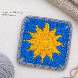 May include: A square crochet piece with a sunburst design in yellow and blue, framed by a gray border. The text "Beginner friendly PDF Pattern" is visible in the upper left corner. The yarn is visible in the background.