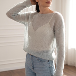Puede incluir: Un suéter de punto de manga larga, transparente y de color azul claro con una textura difusa, usado sobre una camiseta blanca con cuello en V. El suéter presenta una construcción de punto abierto y se combina con jeans de mezclilla azul claro.