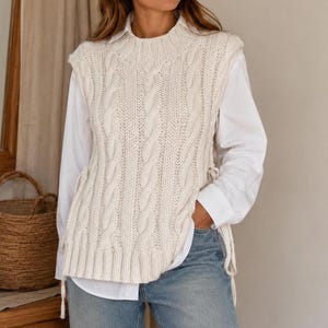 Op de afbeelding: Een crèmekleurig kabelgebreid vest zonder mouwen met een geribbelde ronde hals en zoom, gedragen over een wit overhemd met lange mouwen en een blauwe spijkerbroek. Het vest heeft zijstrikken en een dik, gestructureerd verticaal kabelpatroon.