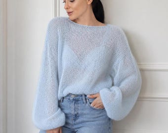 May include: A light blue, loose-knit sweater with a wide boat neckline and long, voluminous sleeves. The sweater features a semi-sheer, airy texture and is paired with light-wash blue denim jeans that have a classic button and zipper fly closure.