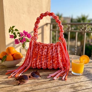 May include: A handmade crochet handbag crafted from thick, braided fabric strips in vibrant shades of orange, pink, and light pink. The bag features a rounded handle and long, dangling fringe tassels on both sides, creating a textured appearance.