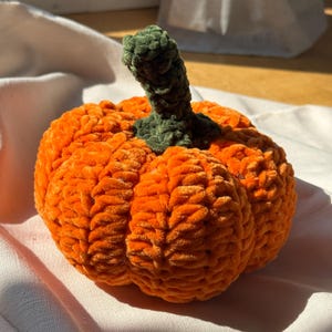 May include: A handcrafted crochet pumpkin made from soft, plush orange yarn with a textured, ribbed surface. The pumpkin features a prominent, dark green crocheted stem extending from the center top, showcasing detailed, looped yarn stitches throughout.
