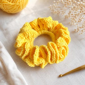 May include: A bright yellow, handmade crochet scrunchie. The scrunchie is made of soft yarn and has a ruffled design. A ball of yellow yarn and a crochet hook are also visible in the image.