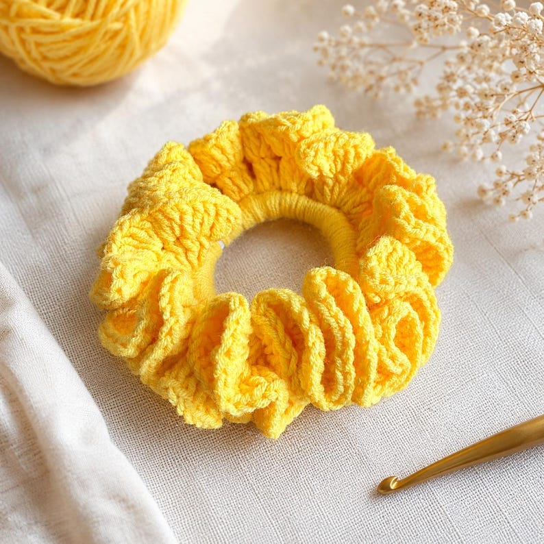 May include: A bright yellow, handmade crochet scrunchie. The scrunchie is made of soft yarn and has a ruffled design. A ball of yellow yarn and a crochet hook are also visible in the image.