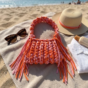 May include: A handmade crochet handbag featuring a vibrant alternating pattern of bright orange and soft pink yarn. The bag includes a thick, braided handle and long, dangling fringe tassels on both sides, crafted from the same orange and pink fabric.