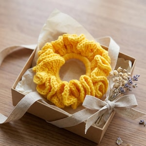 May include: A bright yellow, handmade crochet scrunchie sits in a small, open gift box. The scrunchie is made of thick yarn and has a ruffled design. The box is lined with parchment paper and tied with a beige ribbon. A small bouquet of dried flowers is also included.