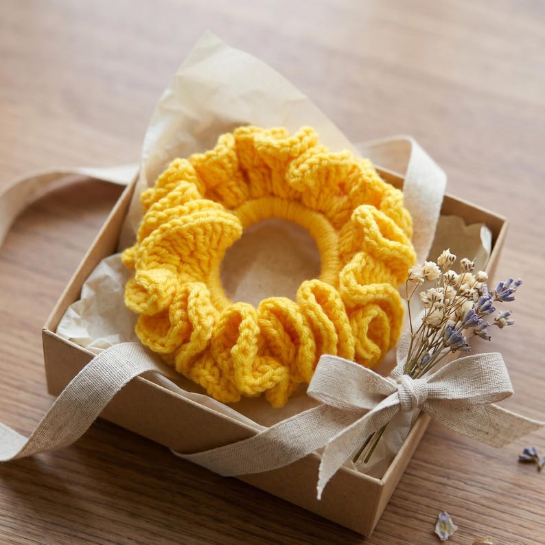 May include: A bright yellow, handmade crochet scrunchie sits in a small, open gift box. The scrunchie is made of thick yarn and has a ruffled design. The box is lined with parchment paper and tied with a beige ribbon. A small bouquet of dried flowers is also included.