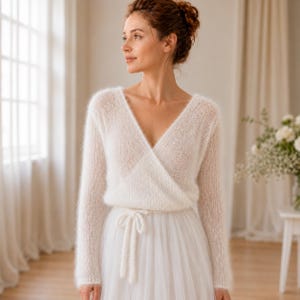 May include: A white long-sleeved wrap sweater made of a soft, fuzzy knit material. The sweater features a deep V-neckline, long sleeves, and a matching tie closure at the waist. The knit texture is light and airy, providing a delicate appearance.