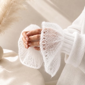 Peut inclure: Un pull blanc à manches longues doté de poignets à volants délicats, transparents et en maille ajourée. Le vêtement est confectionné dans un fil doux et duveteux avec une texture côtelée au poignet, s'évasant en un motif large et aéré.