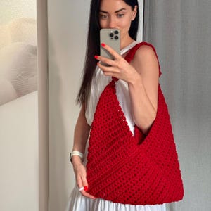 Pode incluir: Uma bolsa de ombro de crochê em vermelho vibrante com um padrão texturizado de pontos abertos. A bolsa apresenta uma silhueta triangular com uma alça larga integrada que descansa sobre o ombro, exibindo uma construção de fio feita à mão.