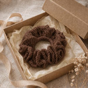 May include: A brown, crocheted scrunchie sits inside an open cardboard box lined with parchment paper. The scrunchie is made of a thick yarn and has a ruffled appearance. A ribbon and dried flowers are also visible.