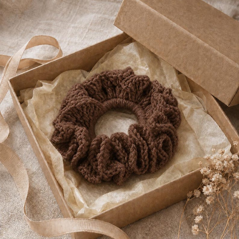 May include: A brown, crocheted scrunchie sits inside an open cardboard box lined with parchment paper. The scrunchie is made of a thick yarn and has a ruffled appearance. A ribbon and dried flowers are also visible.