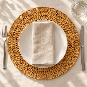 May include: A round mustard yellow crocheted placemat featuring an openwork pattern, topped with a plain white ceramic dinner plate and a folded beige linen napkin. A stainless steel fork and knife are placed on either side of the setting.