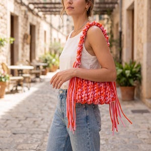 May include: A handmade crochet shoulder bag featuring a chunky knit pattern in vibrant shades of orange, bright pink, and light pink. The bag includes a thick braided strap and long, dangling fringe tassels made from the same colorful yarn strands.