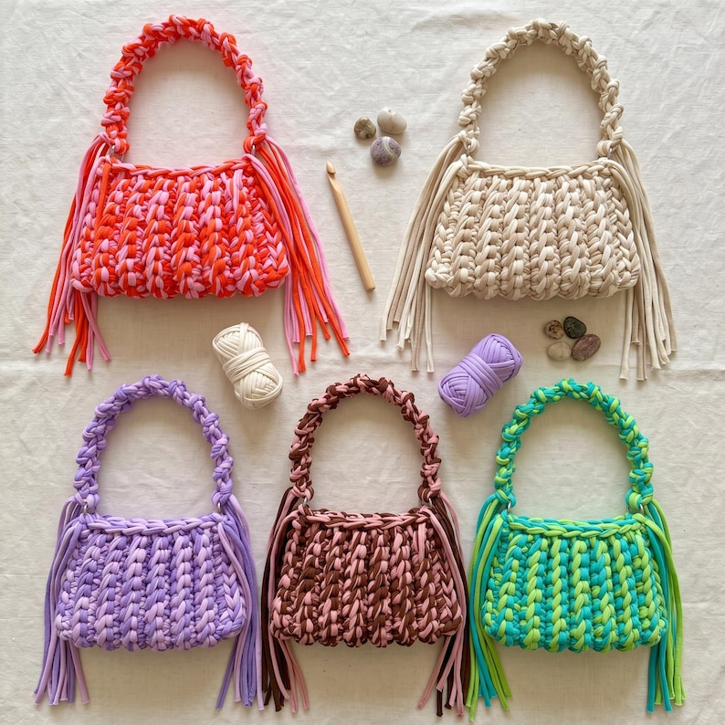 May include: Five handmade crochet handbags with braided handles and long side fringe. The bags are colored in orange and pink, cream, light purple, brown, and a mix of green and yellow. A wooden crochet hook and two small yarn balls are included.