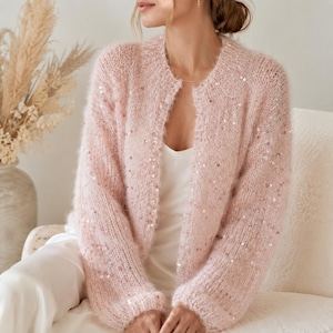 Peut inclure: Un gilet ouvert rose pâle, duveteux, doté de manches longues, d'une encolure ronde et d'un motif dispersé de petites paillettes réfléchissantes sur tout le tricot, porté sur un haut caraco blanc simple.