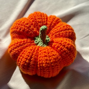 May include: A round, crocheted pumpkin featuring a vibrant orange body with deep, textured ridges and a small, light green stem attached to the center top. The yarn work creates a soft, dimensional appearance with a distinct, hand-crafted finish.