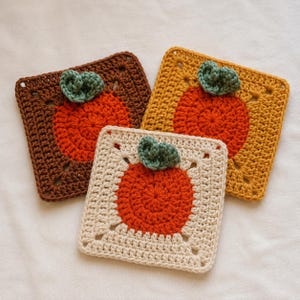 May include: Three square crochet coasters featuring a central orange circular motif and a small green leaf accent. One coaster is dark brown, one is mustard yellow, and one is cream-coloured, all crafted with a textured yarn stitch pattern.