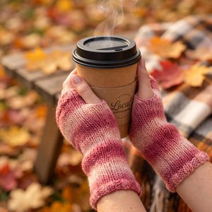Puede incluir: Un par de guantes sin dedos con un patrón de punto jaspeado en tonos rosa y crema. Los guantes se llevan puestos mientras se sujeta un vaso de café de papel color canela con tapa negra y una funda con el texto Local Coffee Shop.