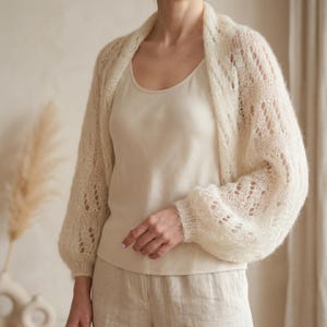 Op de afbeelding: Crèmekleurig gebreid vest met open voorkant en lange, licht gezwollen mouwen. Het vest heeft een opengewerkt ontwerp en wordt gedragen over een crèmekleurige tanktop en beige broek. De stijl is zacht en luchtig.