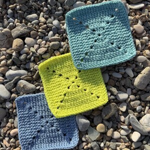 Peut inclure: Trois carr&eacute;s crochet&eacute;s dans des tons de bleu et de jaune sont dispos&eacute;s sur un lit de galets. Les carr&eacute;s pr&eacute;sentent un motif de point textur&eacute; avec un motif central. Les couleurs comprennent un bleu clair, un jaune vif et un bleu plus fonc&eacute;.