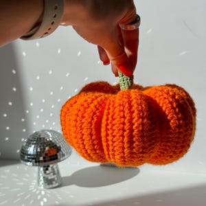 May include: A bright orange crocheted pumpkin with a textured, ribbed surface and a small, light green crocheted stem. The pumpkin is round and plump, featuring distinct vertical segments created by the yarn stitching pattern throughout the piece.