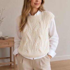 May include: A cream-colored cable knit sweater vest featuring a thick, textured braided pattern across the front. The sleeveless knit garment is layered over a crisp white long-sleeved button-down shirt and paired with light beige trousers.