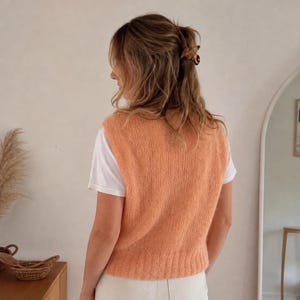 May include: A peach-colored knitted vest worn over a white t-shirt. The vest has a soft, textured appearance. The person is wearing a hair clip. The background includes a mirror and a wooden cabinet.