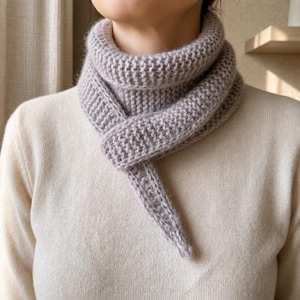 Könnte beinhalten: Hellgrauer Strickschal mit strukturierter Optik. Der Schal ist um den Hals gewickelt und hat ein langes, spitzes Ende. Das Strickmaterial wirkt weich und warm, geeignet für kaltes Wetter.