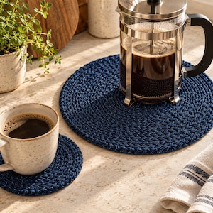 Op de afbeelding: Een ronde placemat van donkerblauw gevlochten touw en een bijpassende kleinere ronde onderzetter. Op de placemat staat een glazen cafetière gevuld met zwarte koffie, terwijl op de onderzetter een gespikkelde keramische mok met koffie staat.
