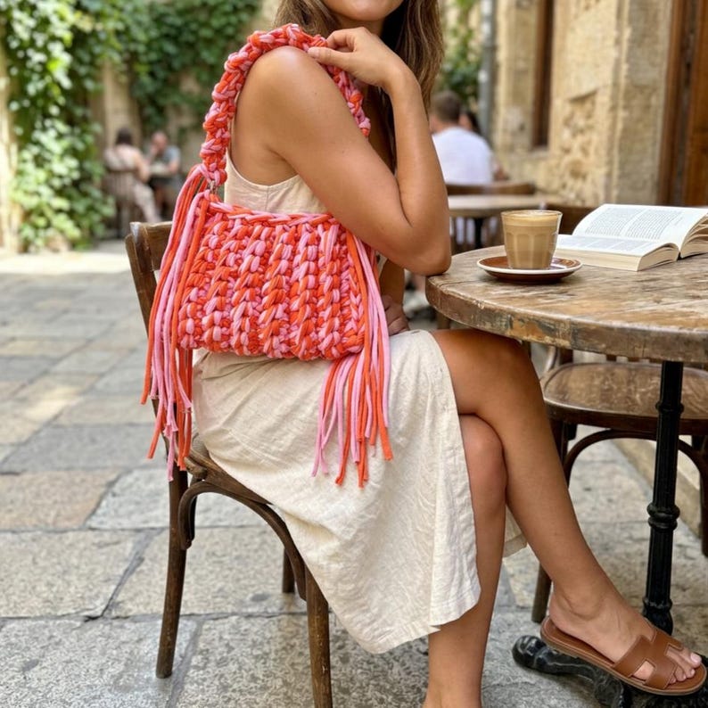 May include: A handmade crochet handbag featuring a vibrant color palette of bright orange and soft pink yarn. The bag has a thick, braided handle and long, dangling fringe tassels hanging from the bottom edges, creating a textured appearance.