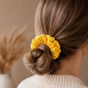 May include: A bright yellow, crocheted scrunchie is worn in a bun. The scrunchie is made of a textured yarn and adds a pop of color. The hair accessory is a stylish addition to any hairstyle.