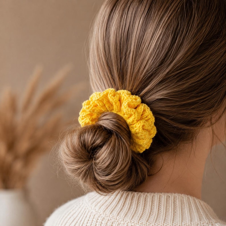 May include: A bright yellow, crocheted scrunchie is worn in a bun. The scrunchie is made of a textured yarn and adds a pop of color. The hair accessory is a stylish addition to any hairstyle.