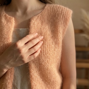May include: A peach-colored, sleeveless knitted vest with a soft, fuzzy texture. The vest has a round neckline and a slightly open front. The material appears to be a delicate, lightweight yarn, perfect for layering. The overall style is simple and elegant.