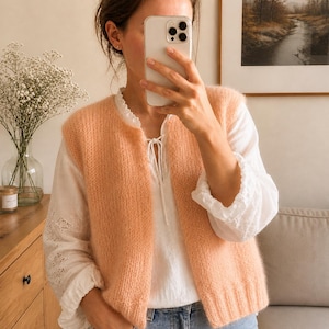 May include: A peach-colored, sleeveless knit vest with a round neckline. The vest is worn over a white blouse with lace detailing on the sleeves and neckline. The person is holding a smartphone in front of their face.