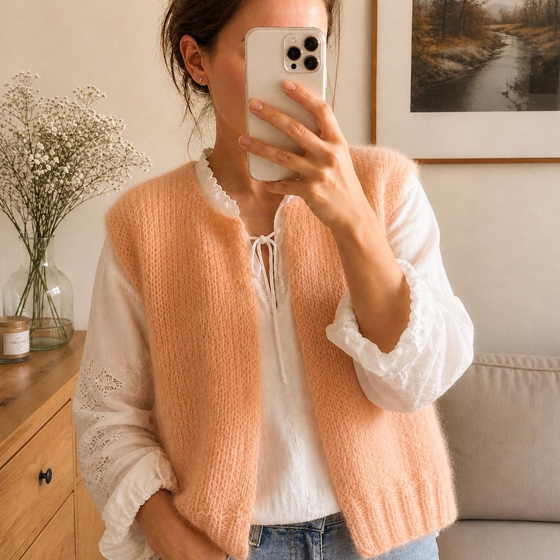 May include: A peach-colored, sleeveless knit vest with a round neckline. The vest is worn over a white blouse with lace detailing on the sleeves and neckline. The person is holding a smartphone in front of their face.