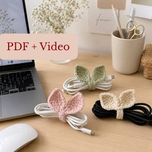 May include: Three handmade crochet cable organizers shaped like bunny ears in sage green, soft pink, and cream colors, each securing a coiled charging cord. A rectangular overlay with the text PDF + Video is positioned in the upper left corner.