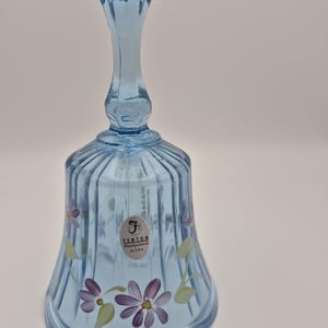 May include: A light blue Fenton hand-painted glass bell with a long handle. The bell features vertical ridges and is decorated with purple and white flowers and green leaves. The bell has a sticker that reads "Fenton Handpainted in USA."