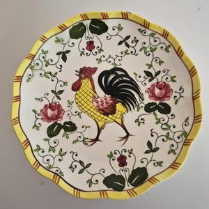 May include: A decorative ceramic plate with a scalloped edge and a yellow border. The plate features a rooster design in yellow, black, and brown, surrounded by floral and vine motifs. The background is white.