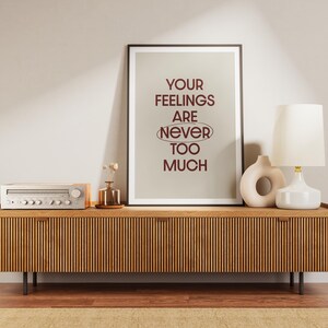 Può includere: Una stampa artistica incorniciata con sfondo beige chiaro e testo rosso scuro che recita: YOUR FEELINGS ARE NEVER TOO MUCH, con la parola NEVER racchiusa in un ovale. La stampa è esposta in una sottile cornice nera su una superficie in legno.